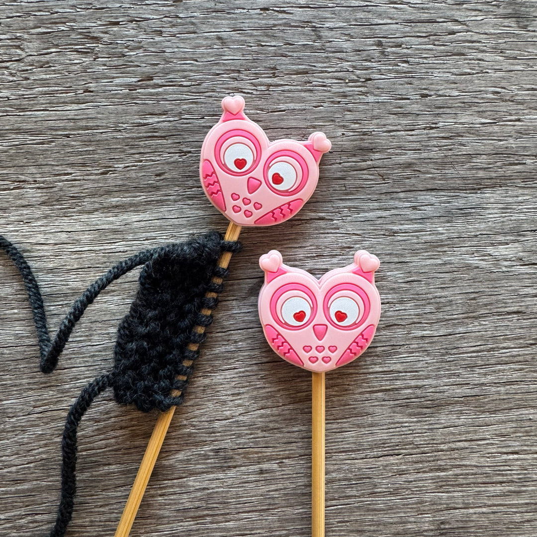 Two pink owl-shaped stitch stoppers on knitting needles with a black yarn ball on a wooden surface.