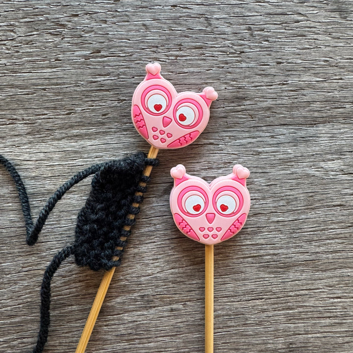 Two pink owl-shaped stitch stoppers on knitting needles with a black yarn ball on a wooden surface.