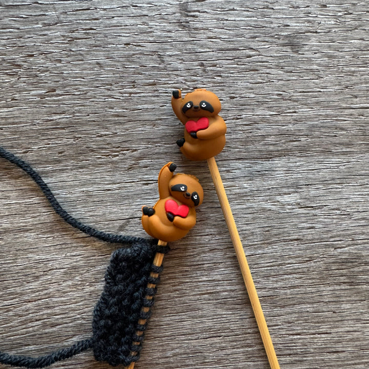Two sloth-shaped stitch stoppers on wooden needles.