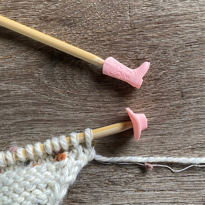 A knitting needle with a pink silicone stitch stopper inserted at the tip, preventing stitches from falling off. There is also an ongoing knitting project in the background.